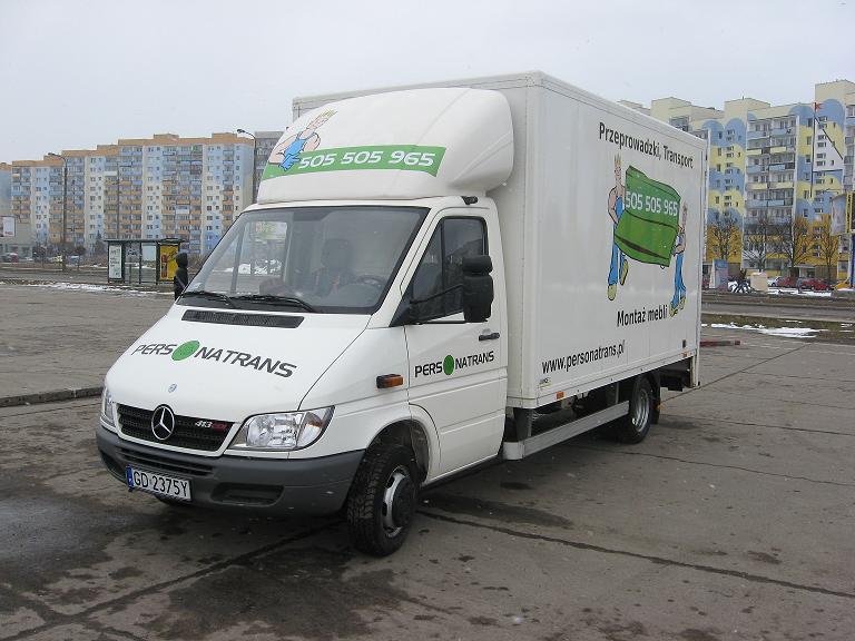 Biały samochód dostawczy marki Mercedes-Benz z logo 'PERSONATRANS' i numerem telefonu 505 505 965 na karoserii, zaparkowany na placu z blokami mieszkalnymi w tle.