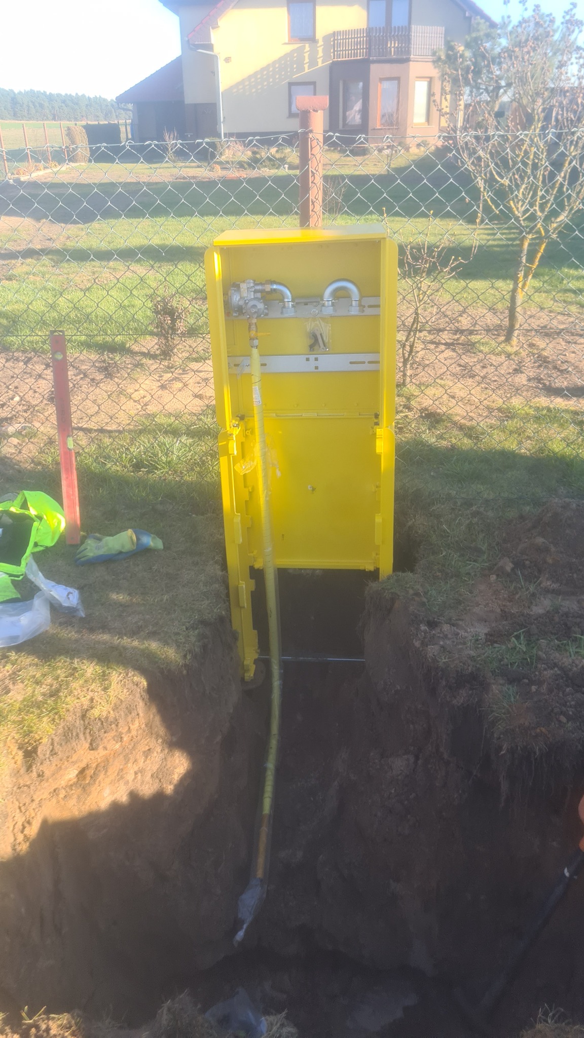 Żółta skrzynka gazowa z otwartymi drzwiczkami, wystająca z wykopu w ziemi, na tle domu jednorodzinnego i ogrodzenia z siatki.