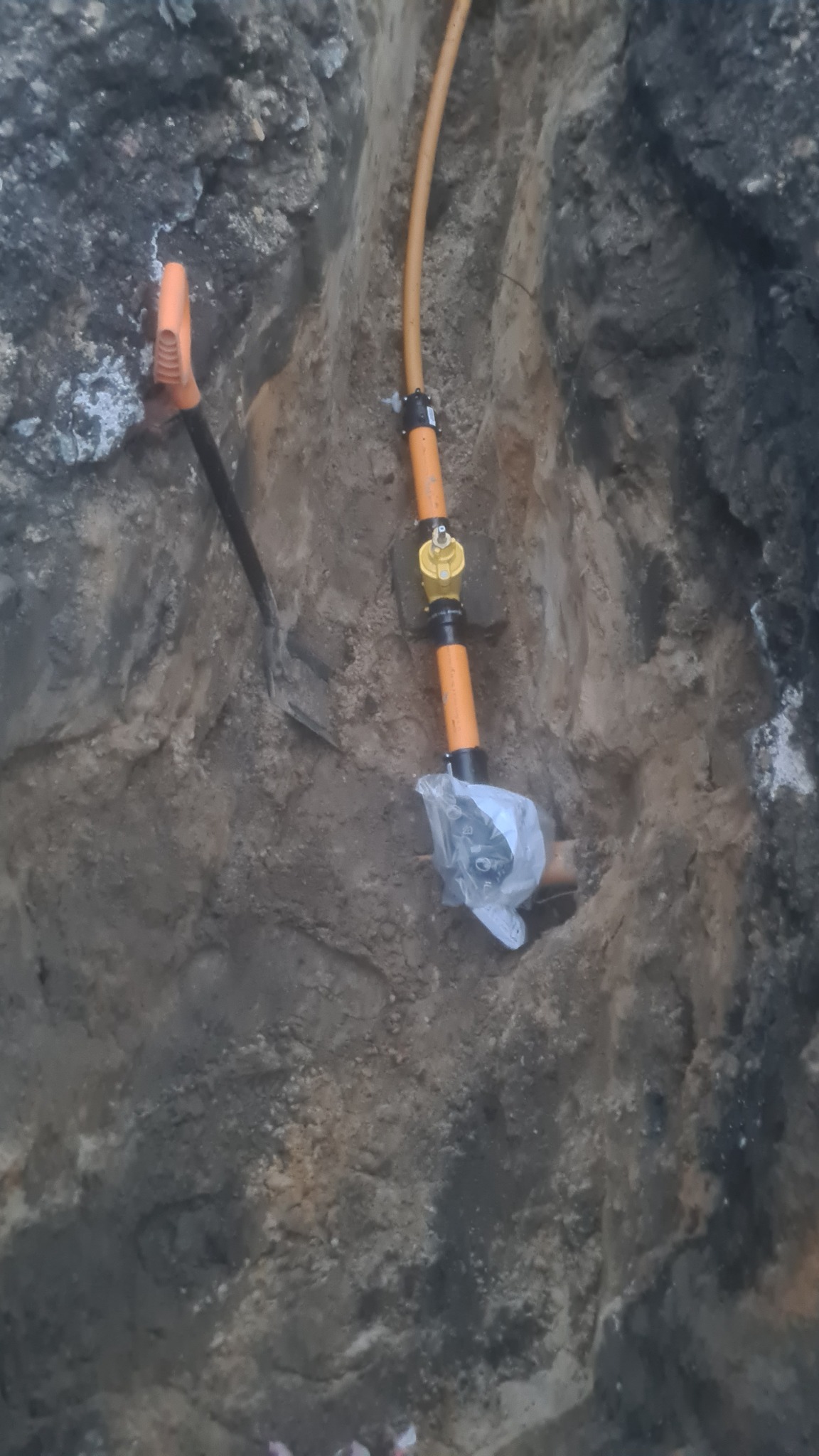 Pomarańczowa rura gazowa z żółtym zaworem w wykopie ziemnym, zabezpieczona folią, obok wbita łopata z pomarańczową rączką.