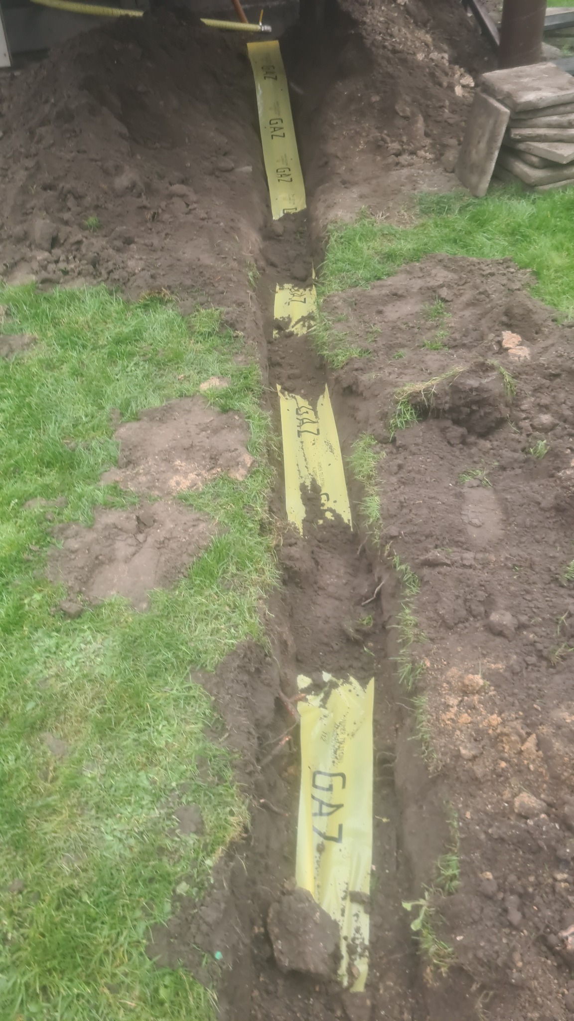 Wykop w ziemi z żółtą folią ostrzegawczą z napisem GAZ, widoczne fragmenty rury gazowej.