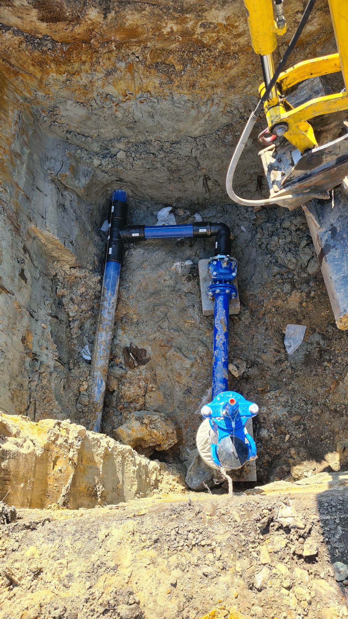 Niebieskie rury wodociągowe z zaworem w wykopie ziemnym, widoczne elementy koparki z żółtymi siłownikami hydraulicznymi.