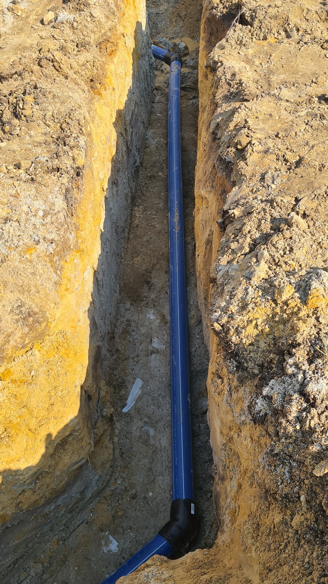 Głęboki wykop w ziemi z ułożoną niebieską rurą wodociągową z czarnymi złączkami, widok z góry.