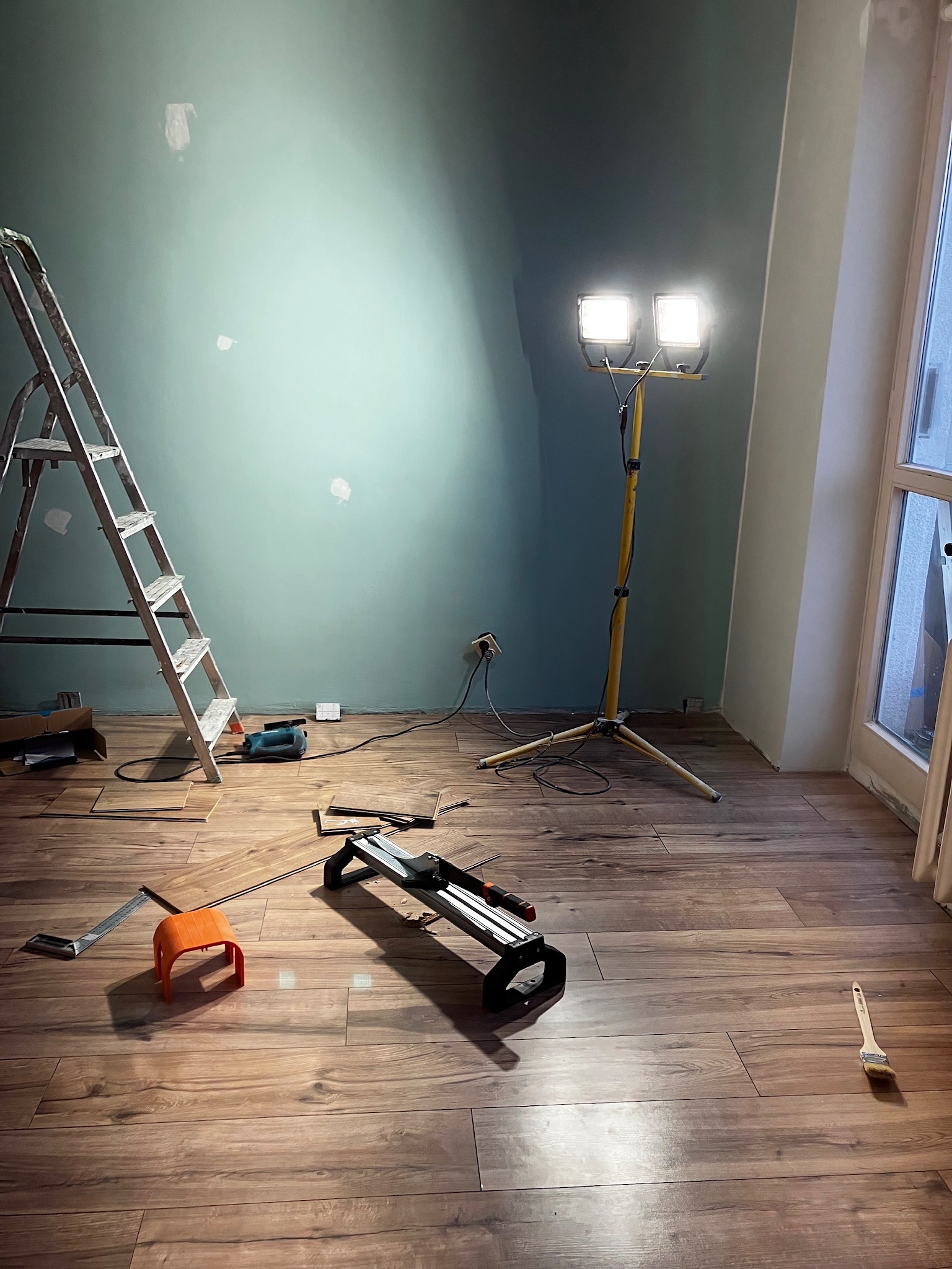 Pokój w trakcie remontu, z niebiesko-zieloną ścianą z widocznymi plamami po szpachlowaniu, drewnianą podłogą, drabiną, narzędziami i lampą budowlaną na statywie.