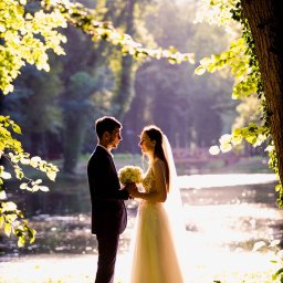 Marcin Klimek Fotograf - Para młoda trzymająca bukiet kwiatów, stojąca w słonecznym parku nad brzegiem stawu, otoczona drzewami z zielonymi liśćmi, w tle widoczny mostek.