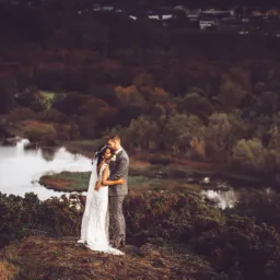 Para młoda obejmuje się na wzgórzu z widokiem na jezioro i las, panna młoda w koronkowej sukni z odkrytymi plecami i wiankiem, pan młody w szarej kracie.