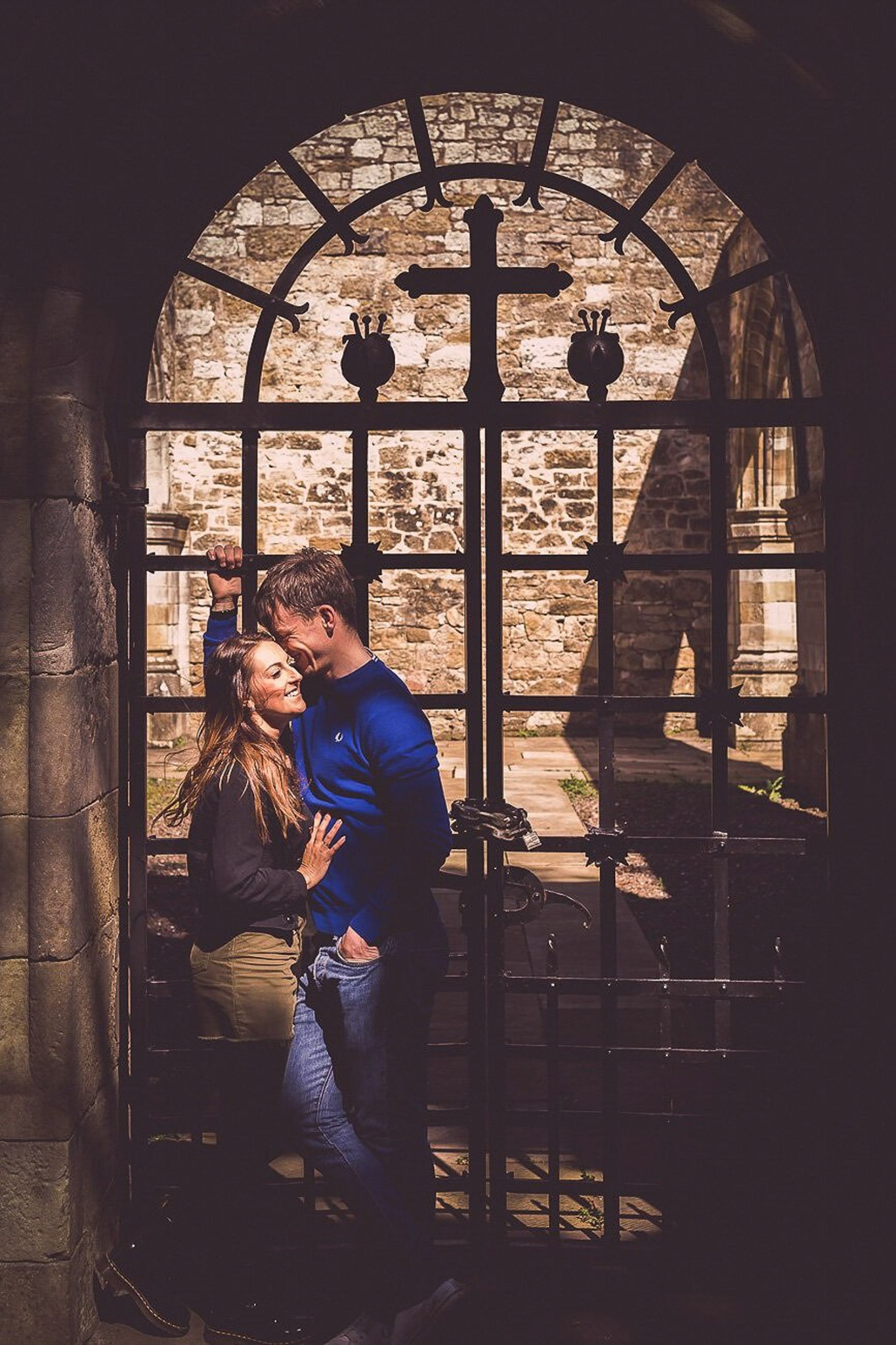Uśmiechnięta para przytulona przy metalowej bramie z krzyżem i dekoracyjnymi elementami na tle kamiennej ściany, delikatne światło pada na ich twarze.