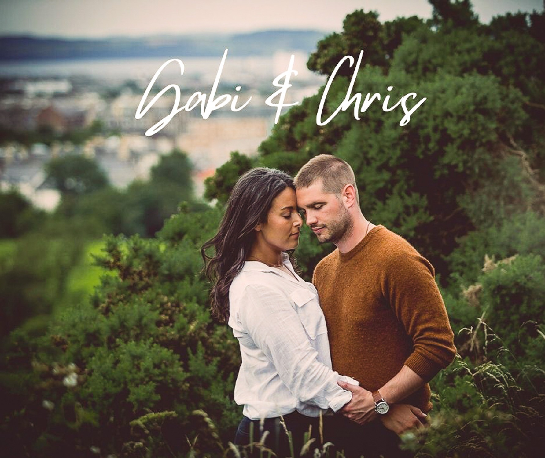 Zakochana para, kobieta w białej koszuli i mężczyzna w brązowym swetrze, obejmują się na tle zieleni z widokiem na miasto Zielona Góra w oddali. Na zdjęciu widnieje napis 'Gabi & Chris'.