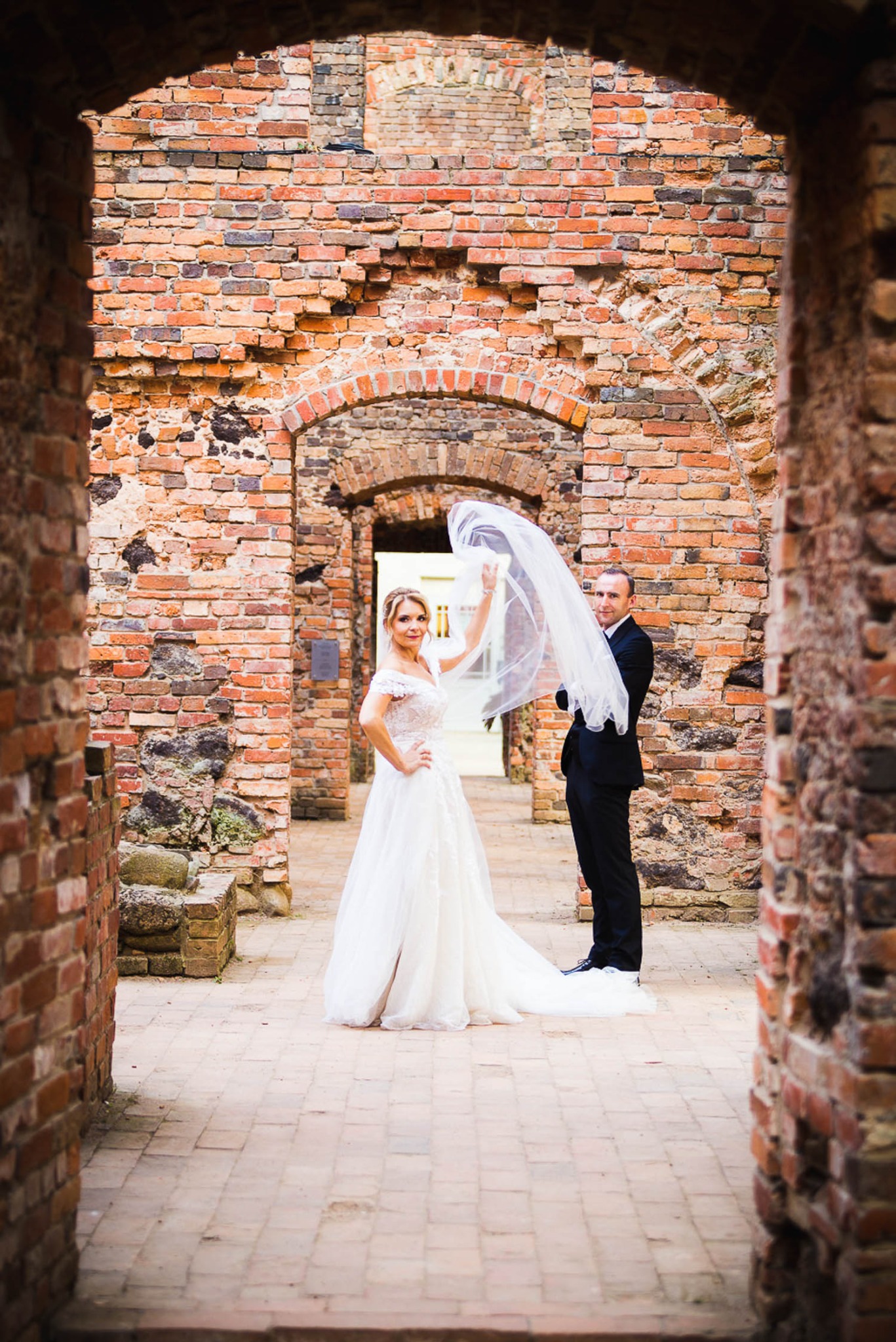 Para młoda pozuje w ruinach ceglanego zamku, panna młoda w koronkowej sukni trzyma welon w górze, pan młody w czarnym garniturze stoi obok.