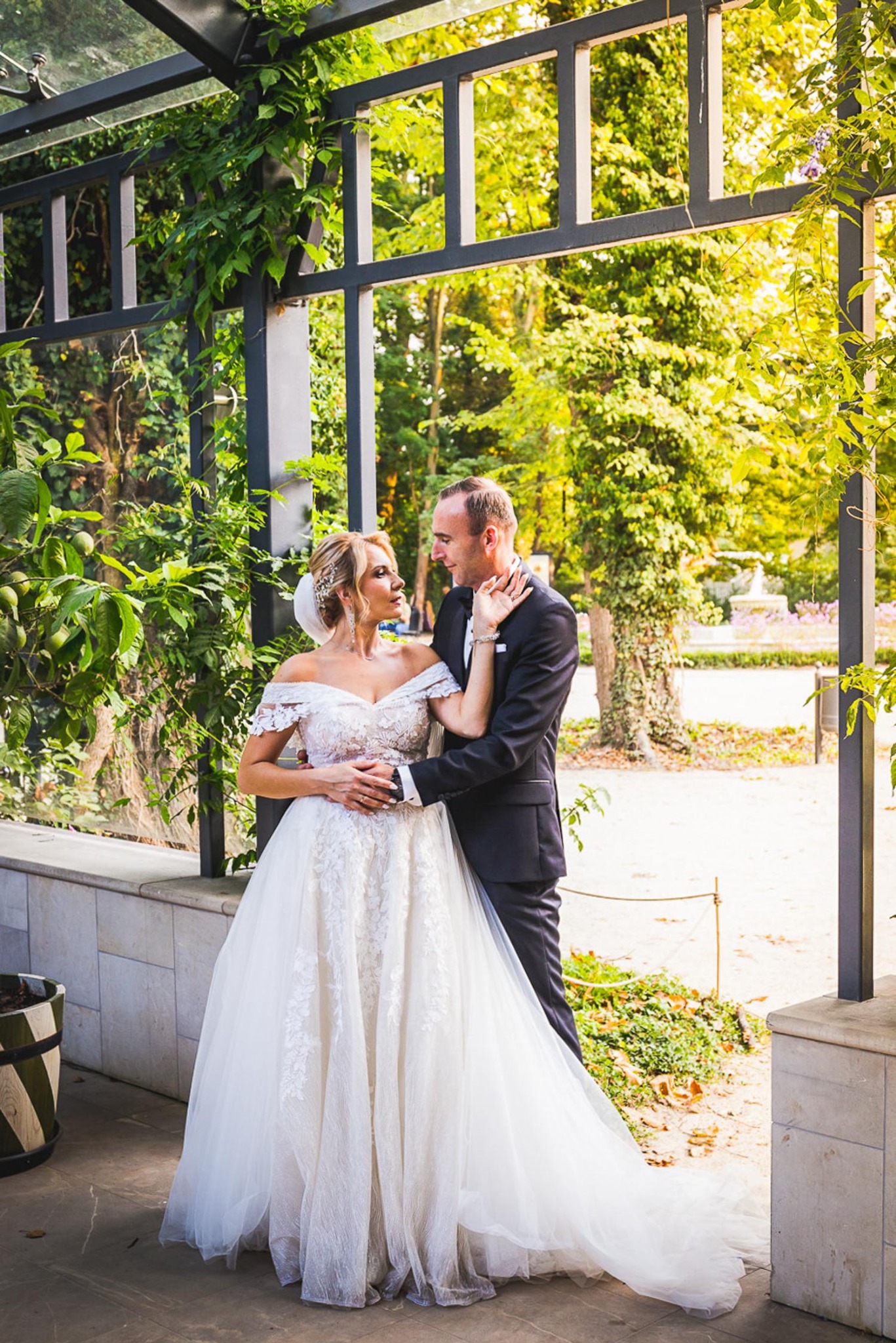 Para młoda w objęciach pod ozdobną pergolą z zielenią, panna młoda w białej sukni z odkrytymi ramionami i upiętymi włosami, pan młody w czarnym garniturze, w tle park z fontanną.