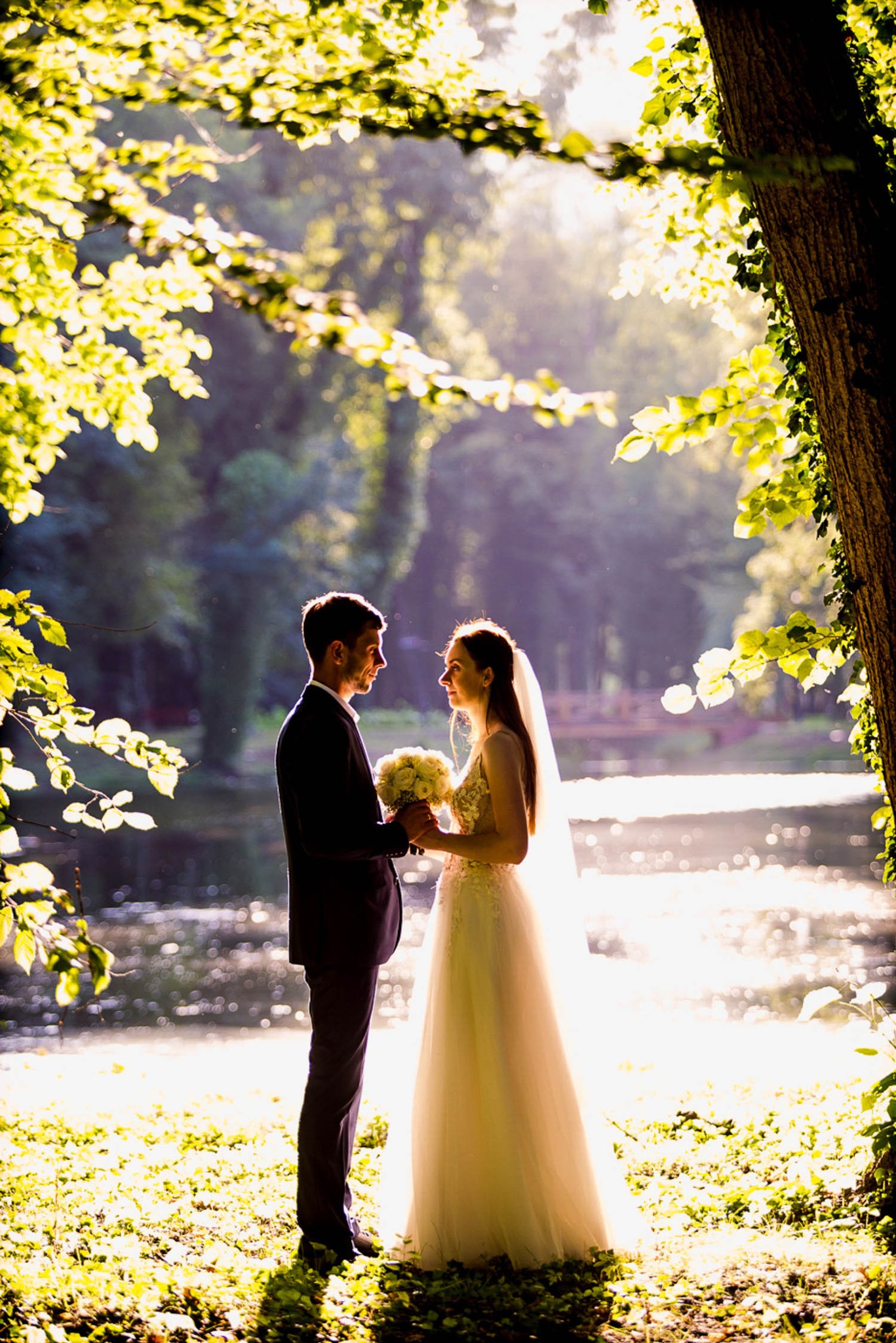 Para młoda trzymająca bukiet kwiatów, stojąca w słonecznym parku nad brzegiem stawu, otoczona drzewami z zielonymi liśćmi, w tle widoczny mostek.