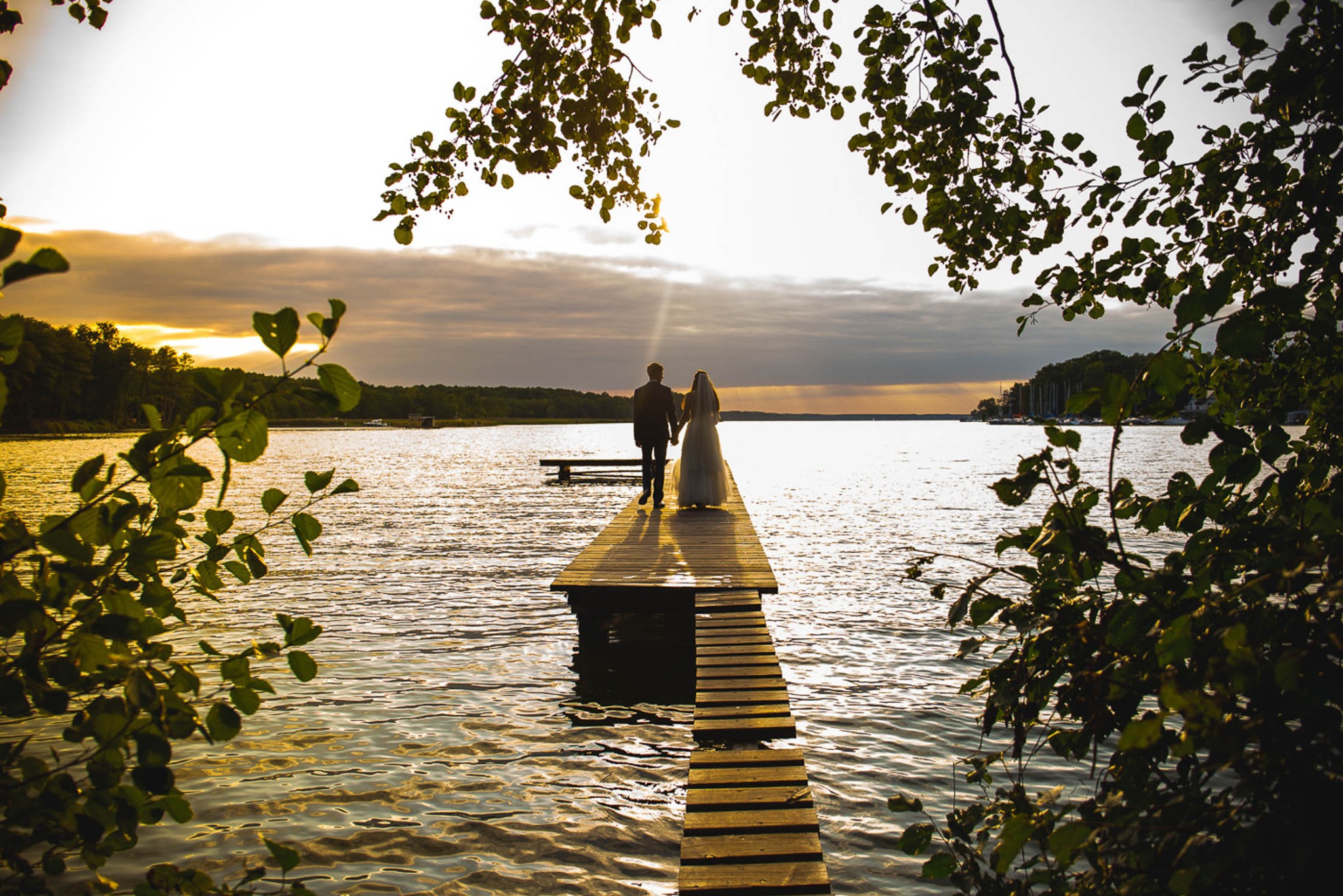 Para młoda idzie drewnianym pomostem w głąb jeziora o zachodzie słońca, widok zza drzew z liśćmi