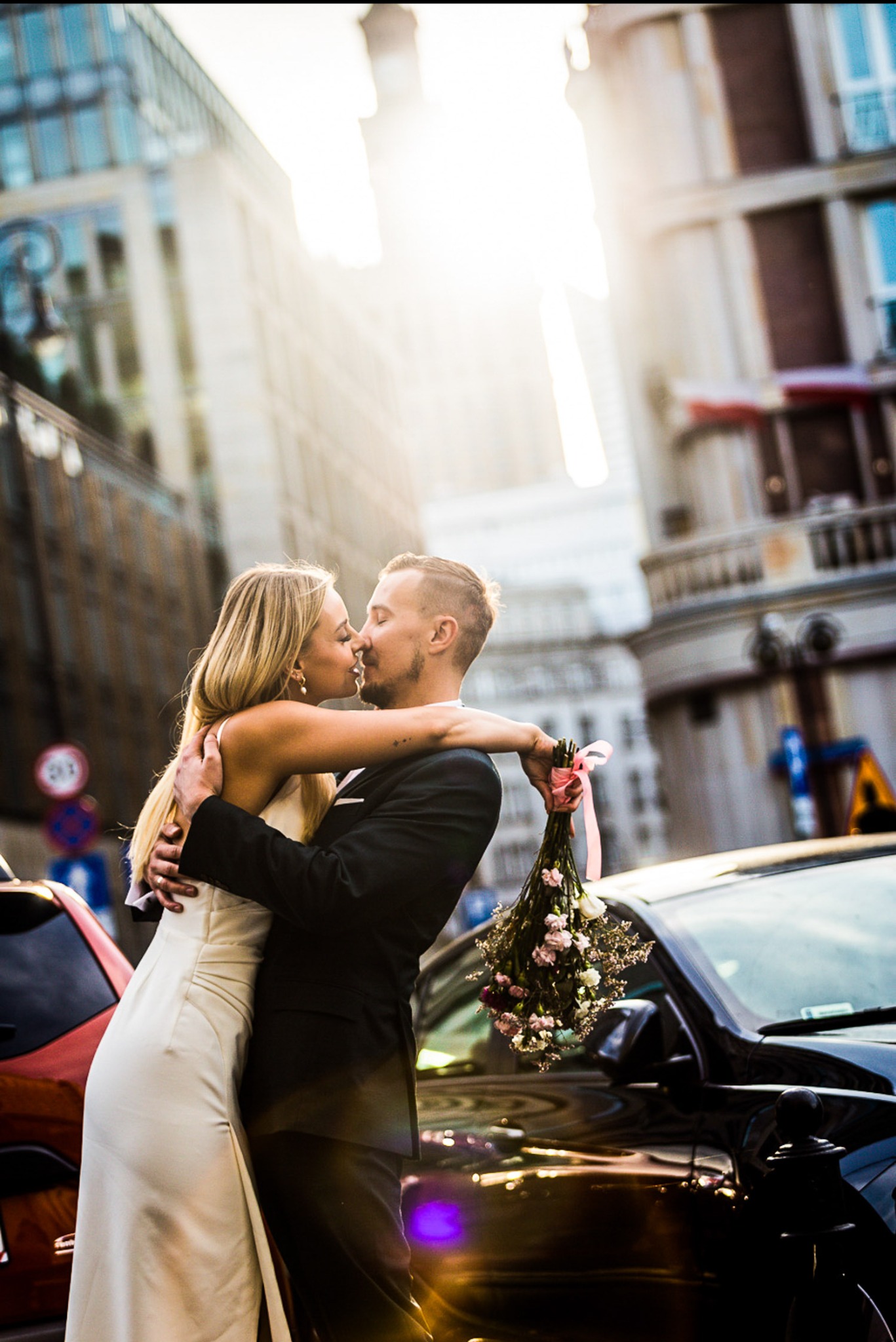 Pocałunek pary młodej na tle miejskiej architektury, bukiet ślubny z różową wstążką, promienie słoneczne.