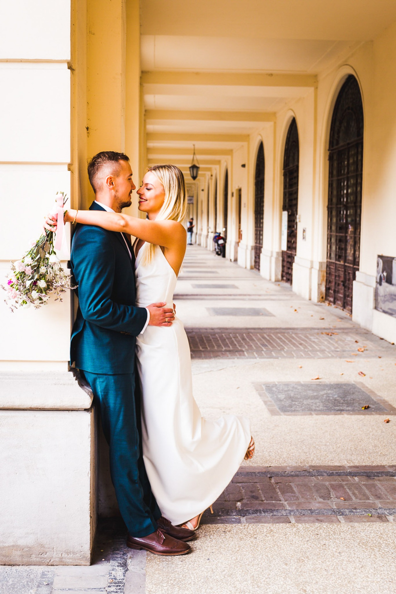 Para młoda w objęciach pod arkadami: panna młoda w prostej białej sukni, pan młody w ciemnozielonym garniturze, bukiet w dłoni, subtelne uśmiechy, ciepłe światło.