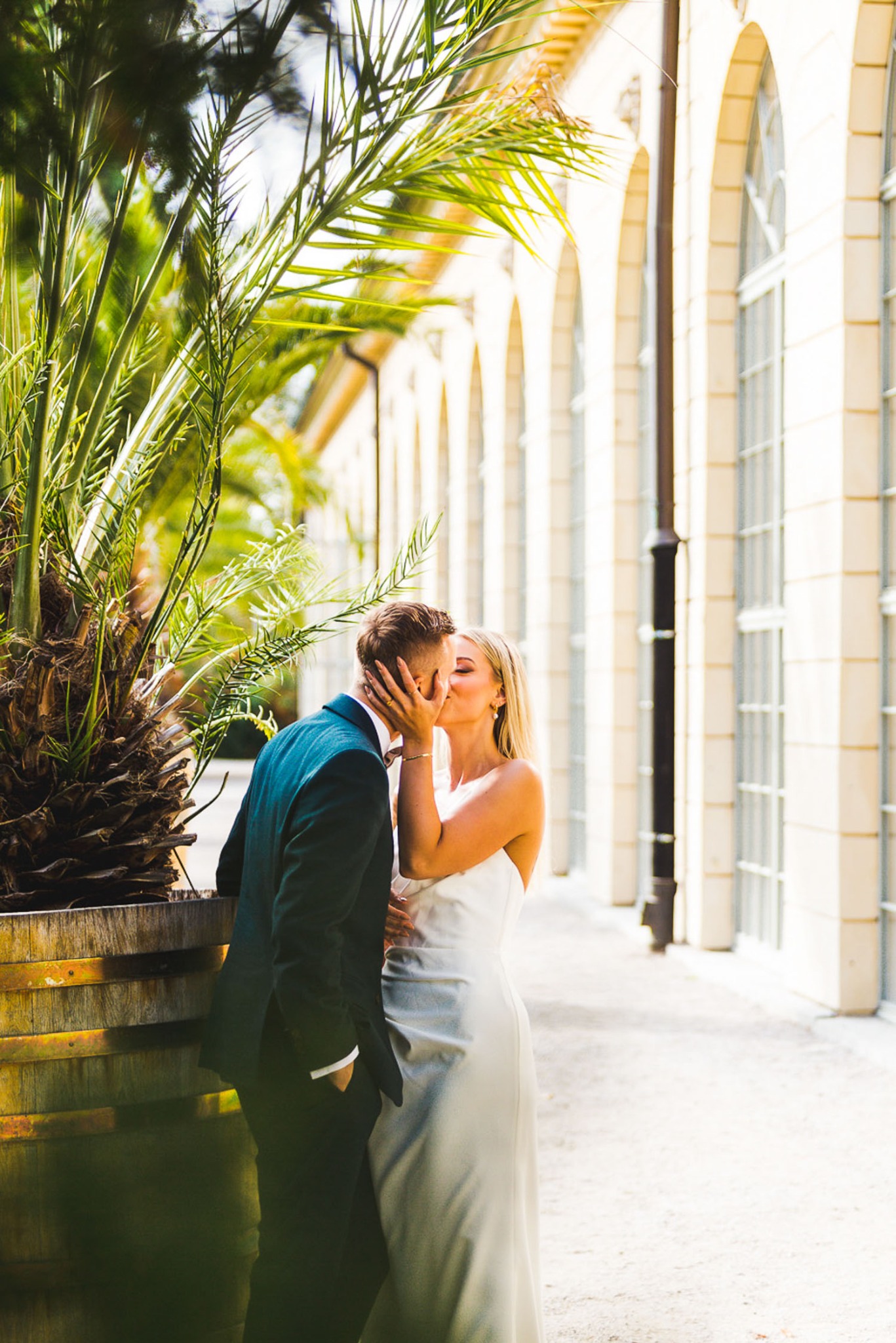 Para młoda w eleganckich strojach całuje się na tle jasnej, klasycznej architektury z palmą po lewej stronie kadru.