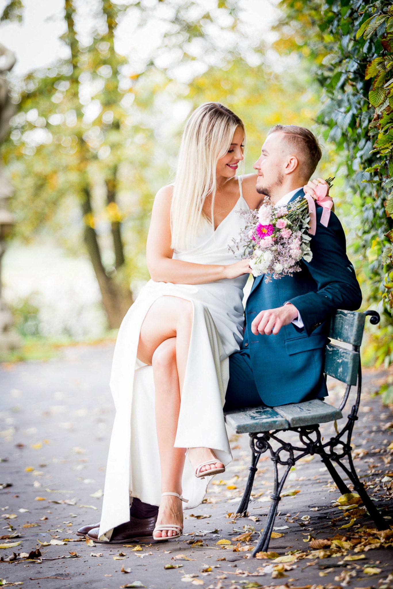 Uśmiechnięta para młoda siedząca na ławce w parku; panna młoda w białej sukni z rozcięciem, pan młody w ciemnozielonym garniturze z bukiecikiem; jesienne liście na ścieżce.