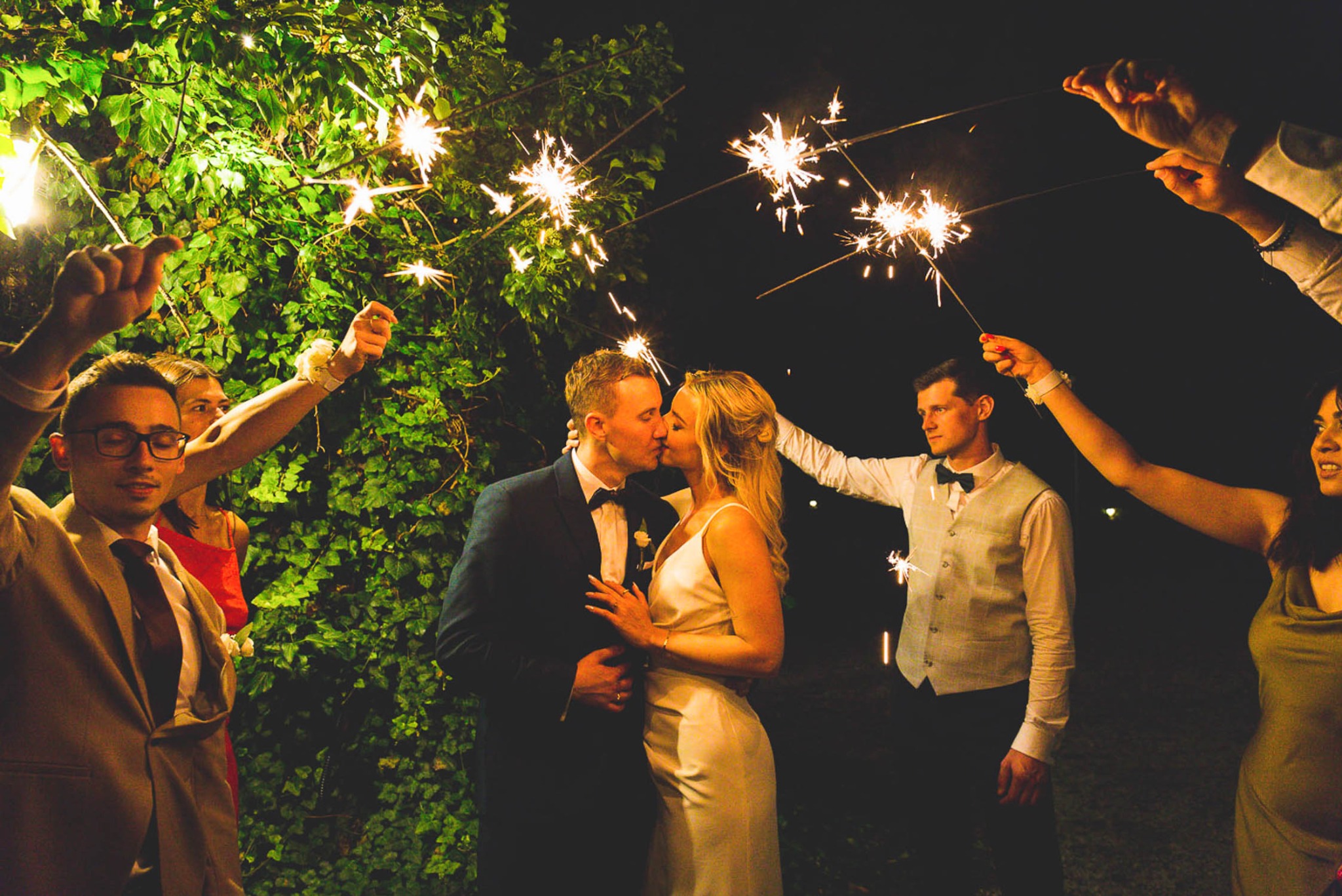 Pocałunek nowożeńców w otoczeniu gości z zimnymi ogniami, nocna sceneria z zieloną roślinnością w tle.