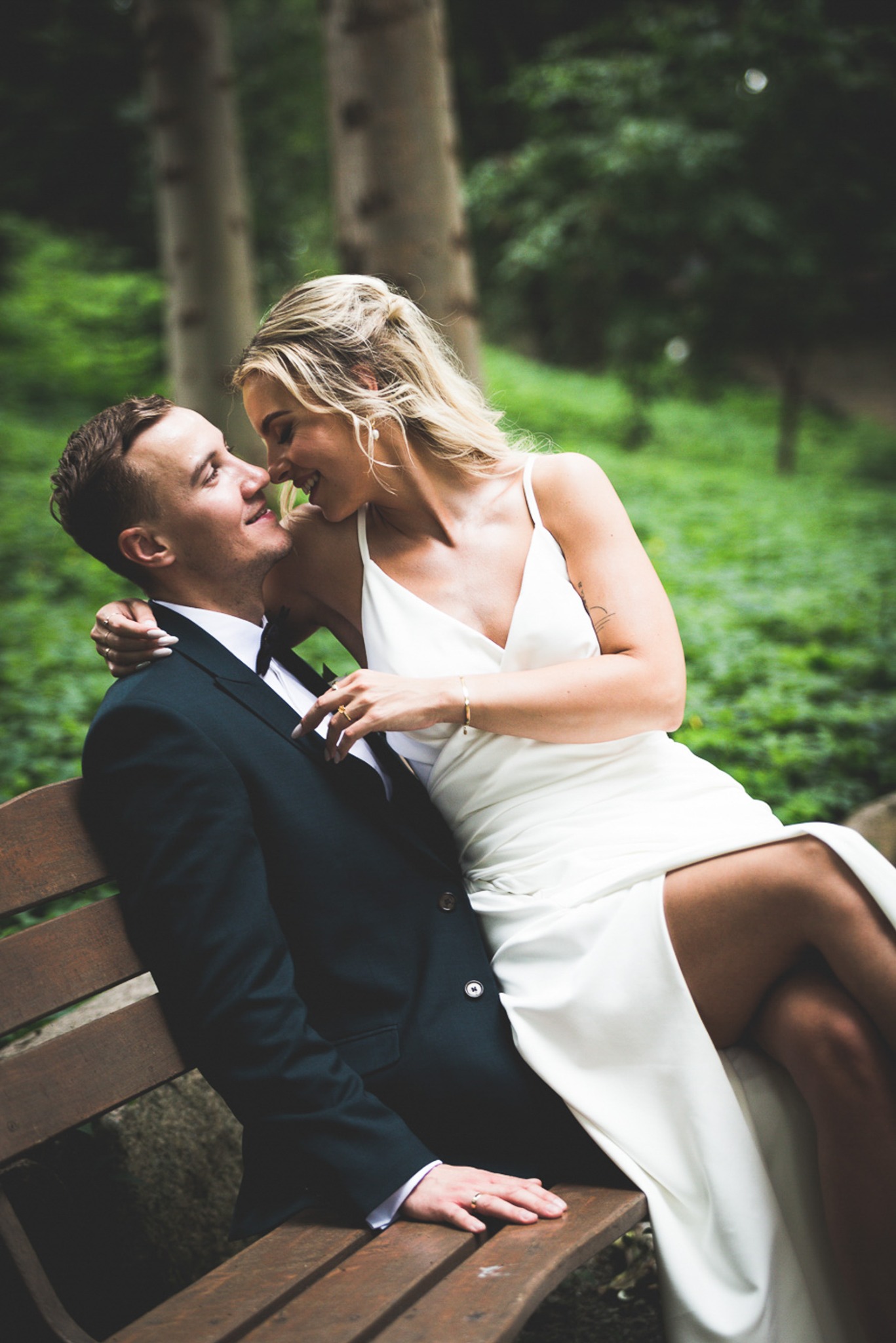 Uśmiechnięta panna młoda w białej sukni z rozcięciem, obejmująca pana młodego w ciemnozielonym smokingu siedzącego na drewnianej ławce w parku, delikatne oświetlenie podkreśla intymną chwilę.