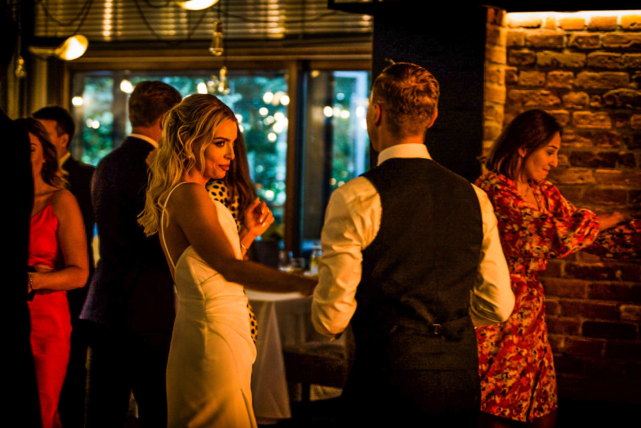 Goście weselni w trakcie zabawy, panna młoda w białej sukni z odkrytymi plecami, pan młody w ciemnej kamizelce, ceglana ściana i okno z dekoracyjnymi lampkami w tle.