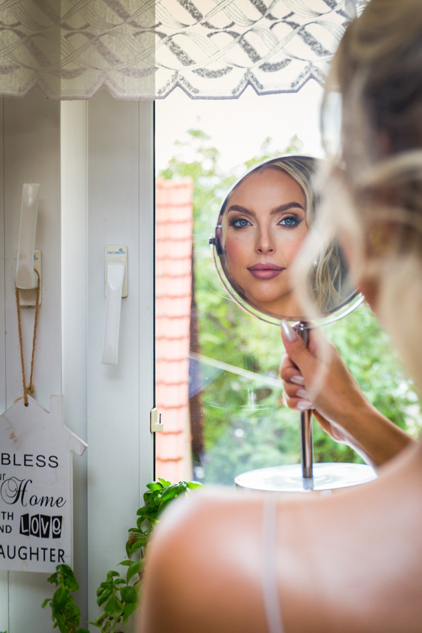 Kobieta przegląda się w okrągłym lusterku, widoczny makijaż z podkreślonymi niebieskimi oczami, jasne włosy, w tle okno z widokiem na zieleń i dekoracyjny napis 'Bless our Home with Love and...
