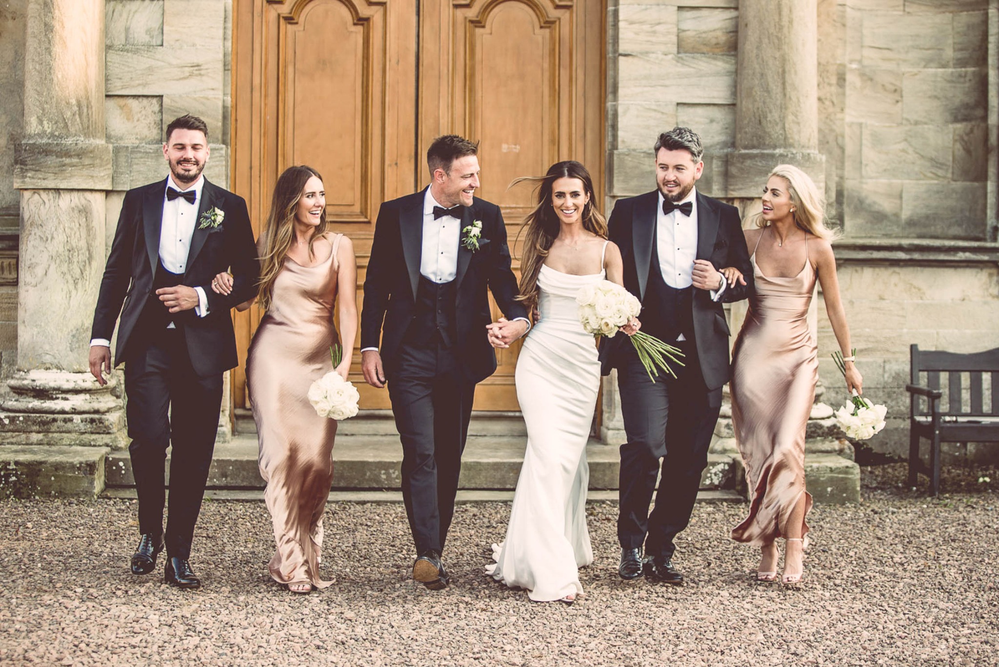 Para młoda w bieli, trzymająca się za ręce, otoczona drużbami i druhnami w smokingach i sukniach w kolorze szampana, spacerująca po żwirowej ścieżce przed budynkiem z kolumnami.