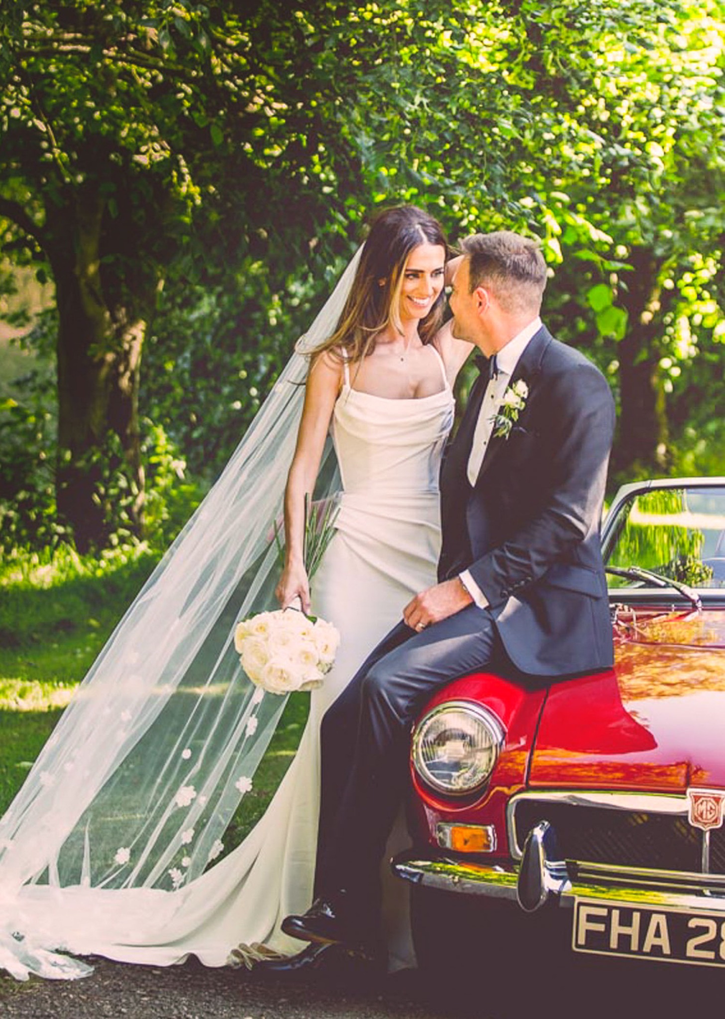 Para młoda: panna młoda w białej sukni i welonie z bukietem, pan młody w czarnym garniturze, siedzący na czerwonym oldtimerze na tle zieleni.