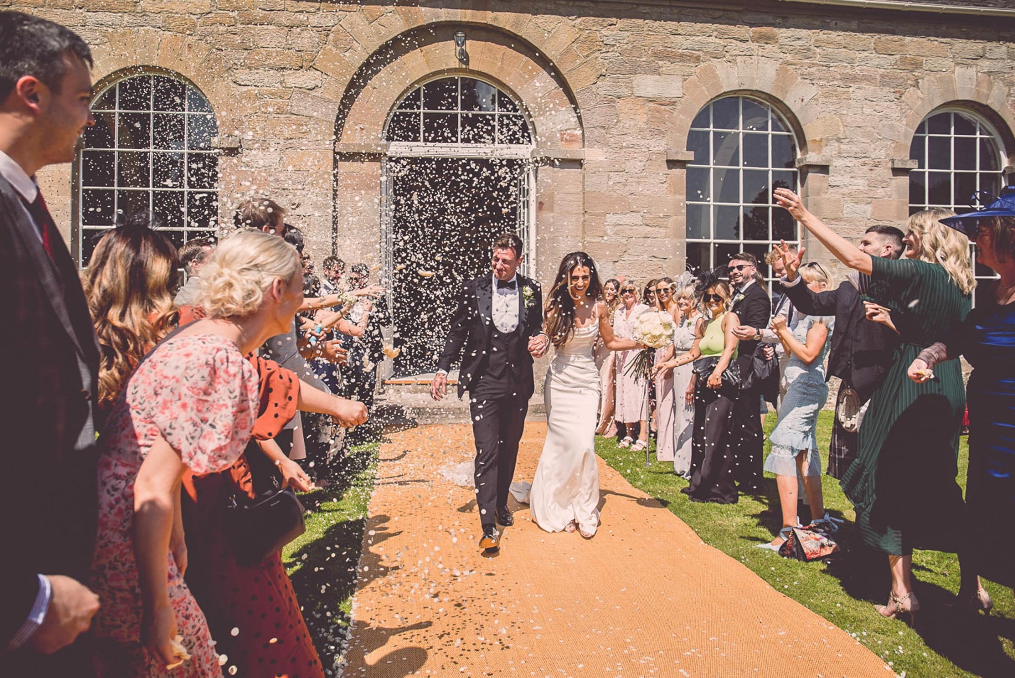 Para młoda wychodzi z kamiennego budynku, obsypywana konfetti przez gości, na tle łukowych okien i pomarańczowego dywanu.