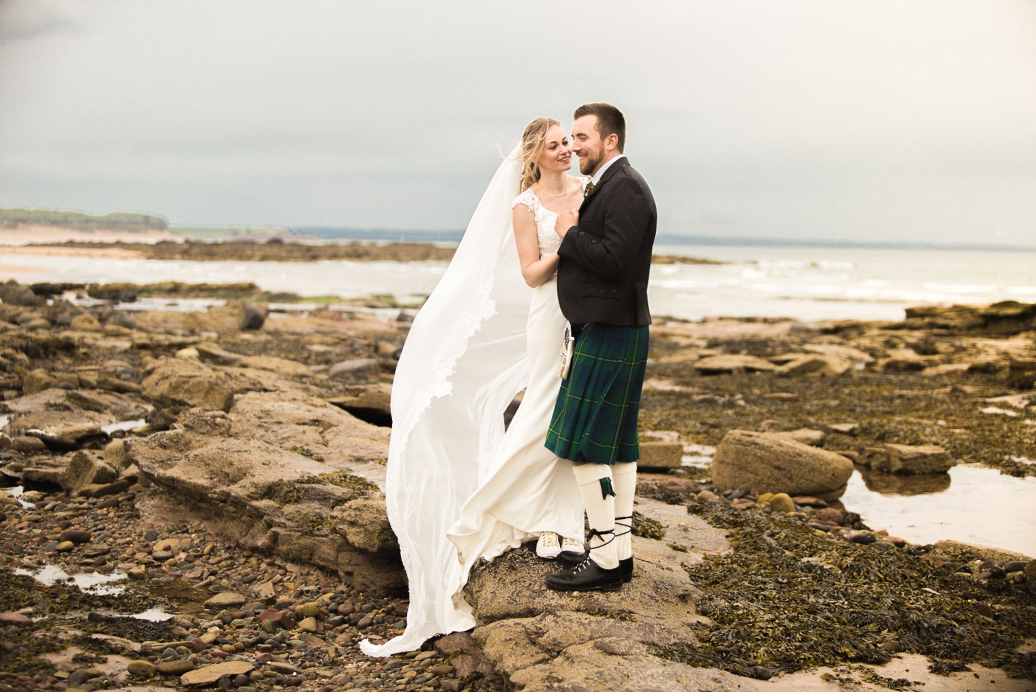 Para młoda, on w szkockim stroju, ona w białej sukni, stoją na skalistym wybrzeżu morza, z czułością patrząc na siebie.
