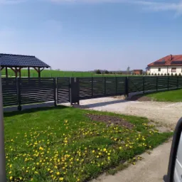 Nowoczesne, poziome, szare ogrodzenie z bramą wjazdową, widok zza lusterka samochodu na tle zielonego krajobrazu i zabudowań mieszkalnych.