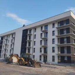 Nowoczesny budynek wielorodzinny z białoszarą elewacją i balkonami, w trakcie prac wykończeniowych. Żółta koparko-ładowarka na pierwszym planie, materiały budowlane obok.