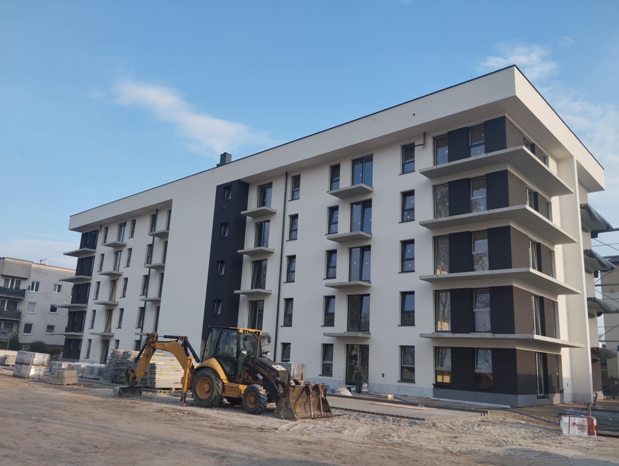 Nowoczesny budynek wielorodzinny z białoszarą elewacją i balkonami, w trakcie prac wykończeniowych. Żółta koparko-ładowarka na pierwszym planie, materiały budowlane obok.