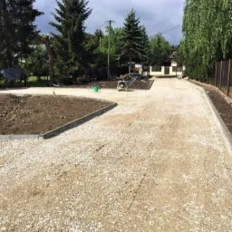 Utwardzona droga żwirowa z obrzeżami betonowymi, świeżo zasiana ziemia po bokach, w tle widoczny dom i drzewa.