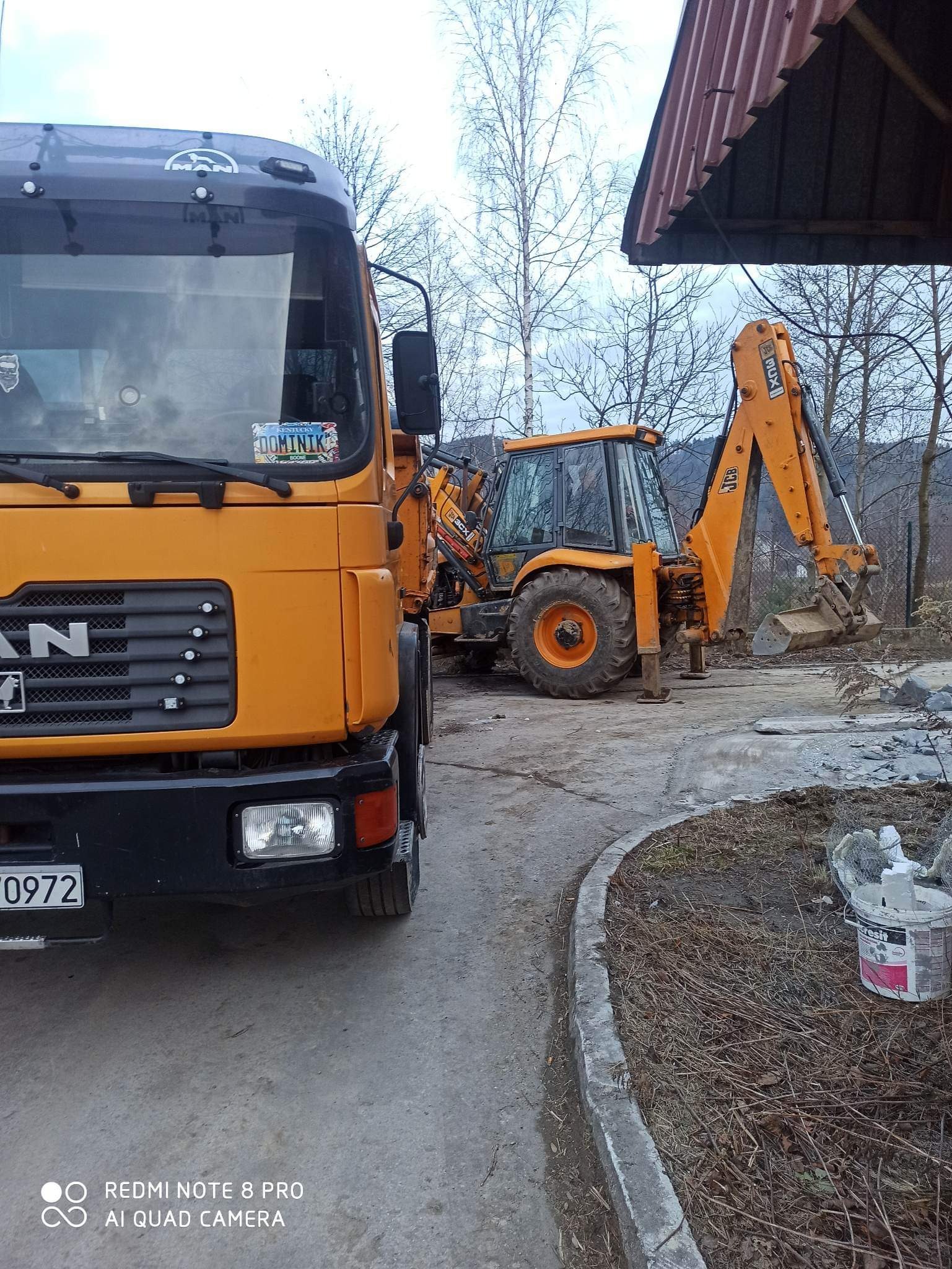 Pomarańczowy samochód ciężarowy MAN z tablicą rejestracyjną Kentucky i koparko-ładowarka JCB 3CX na placu budowy, widoczne drzewa w tle.