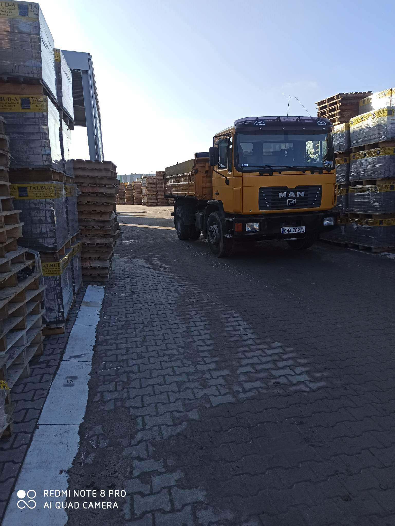 Żółta ciężarówka MAN z otwartą skrzynią ładunkową, zaparkowana na placu wyłożonym kostką brukową, z paletami kostki po bokach. Rejestracja KWA-70972.