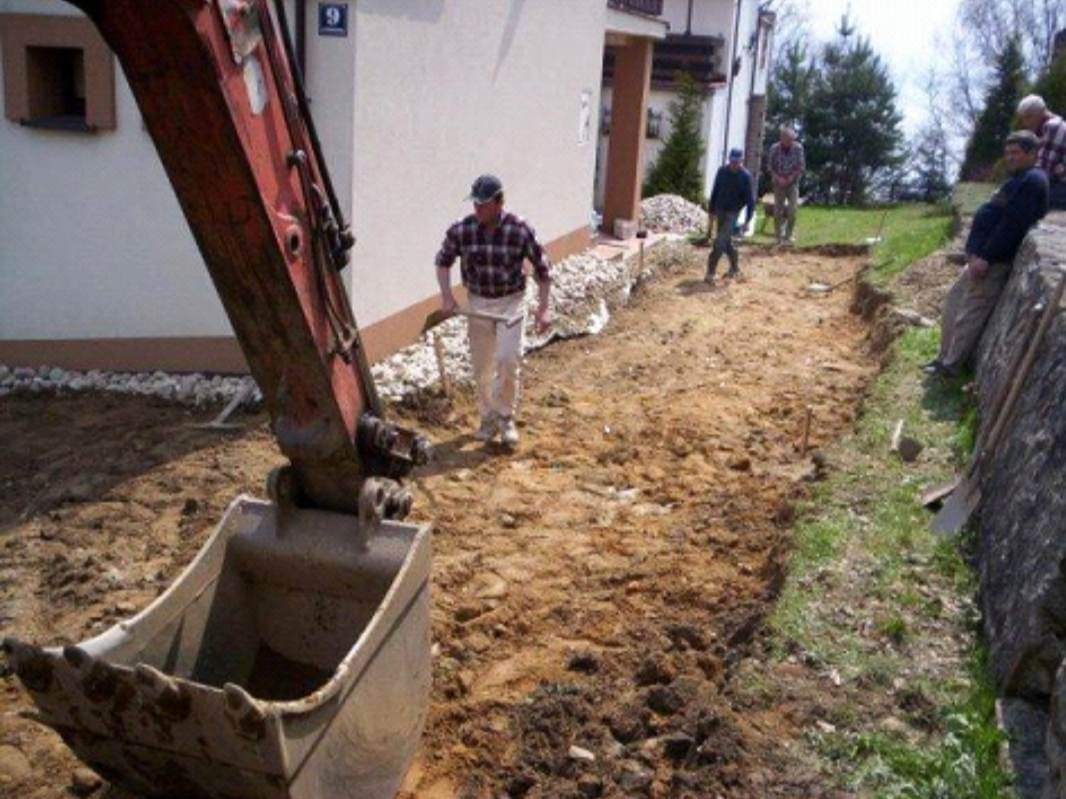 Czerwony wysięgnik koparki z łyżką w trakcie wykopu pod fundamenty domu jednorodzinnego, w tle pracownicy z narzędziami.