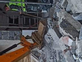 Operator w kamizelce odblaskowej obsługuje koparkę, usuwając gruz po wyburzeniu ściany. Widoczny fragment drabiny i konstrukcji sufitu.