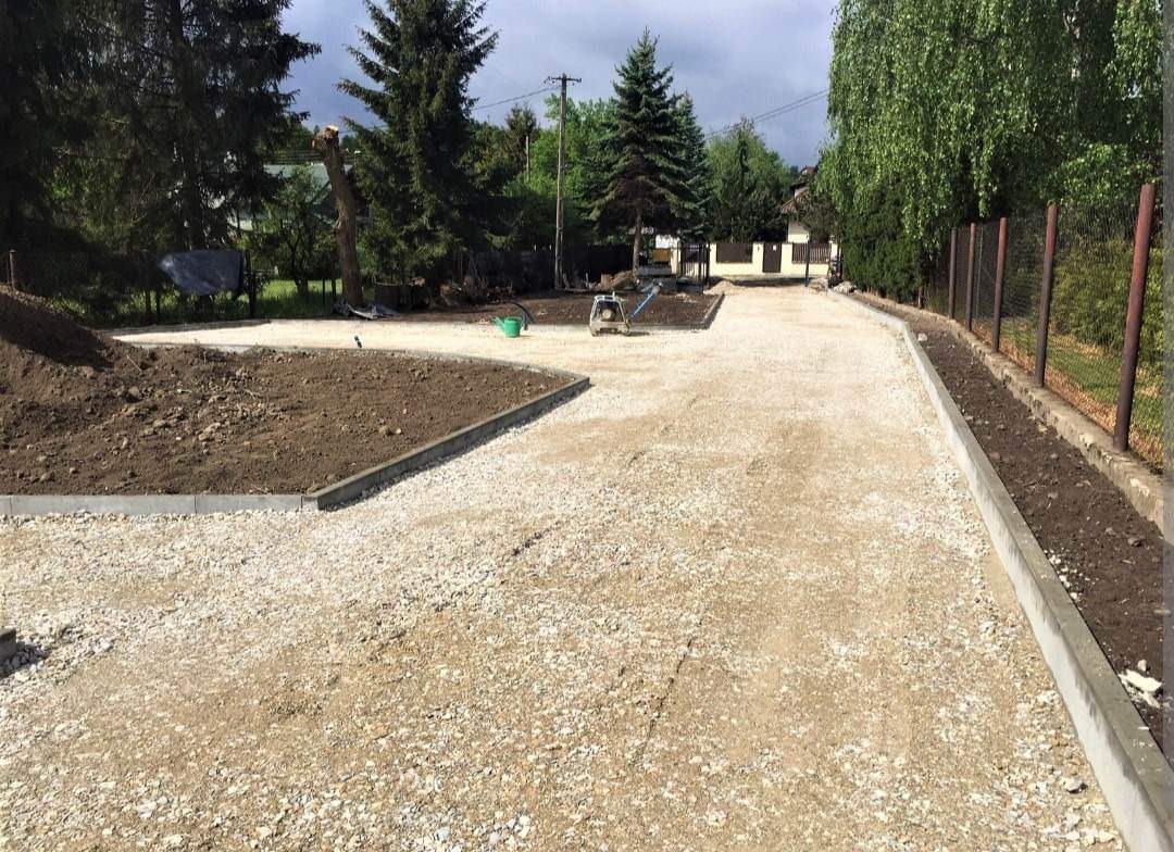 Utwardzona droga żwirowa z obrzeżami betonowymi, świeżo zasiana ziemia po bokach, w tle widoczny dom i drzewa.