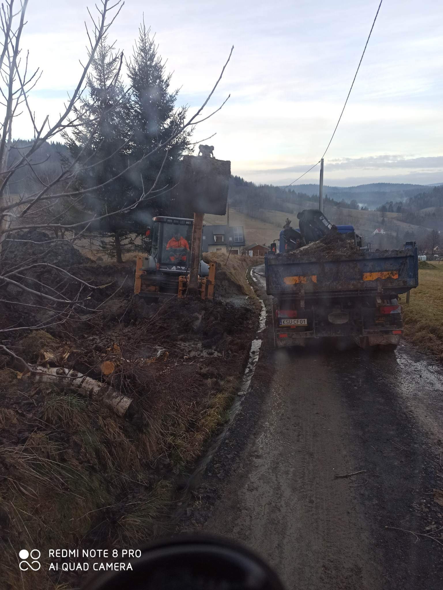 Żółta ładowarka czołowa z operatorem w pomarańczowej kurtce załadowuje ziemię do wywrotki na błotnistej drodze, w tle widoczne wzgórza i drzewa.