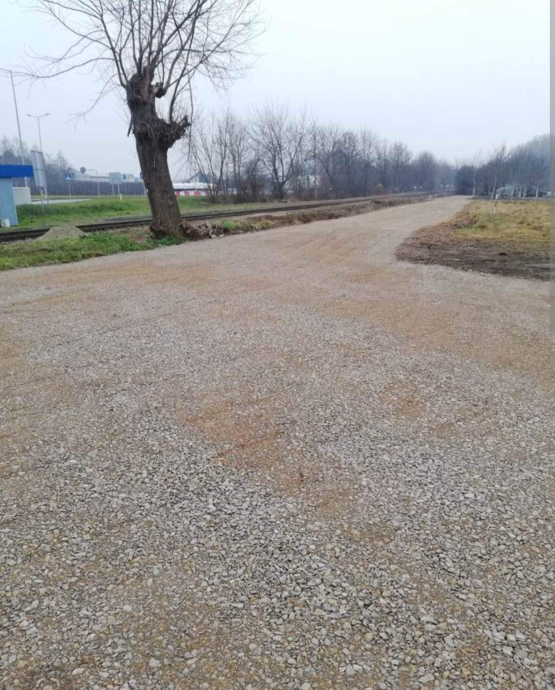 Świeżo wysypana, szutrowa droga gruntowa z widocznym drzewem przy krawędzi i torami kolejowymi w tle, pochmurny dzień.