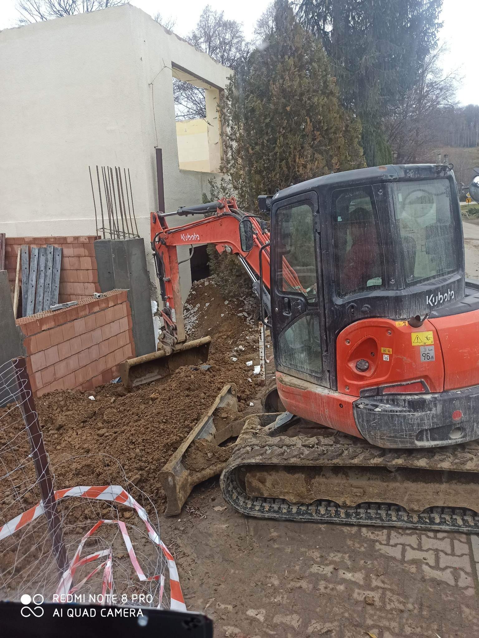 Pomarańczowa minikoparka Kubota podczas prac ziemnych przy częściowo rozebranym budynku, widoczne fundamenty i gruz.