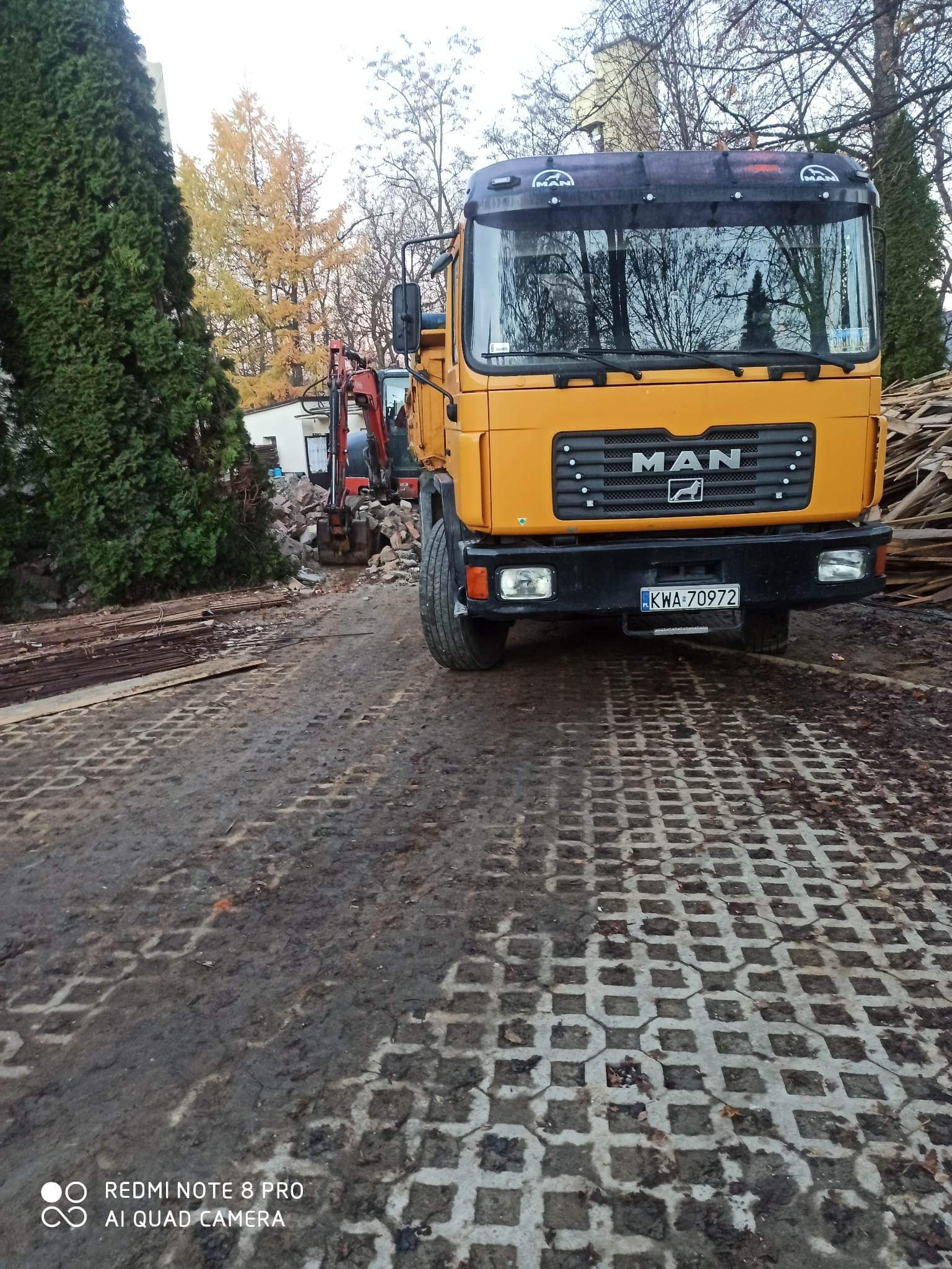 Żółta ciężarówka MAN na placu z kostki ażurowej, w tle minikoparka przy stercie gruzu, prawdopodobnie po wyburzeniu.