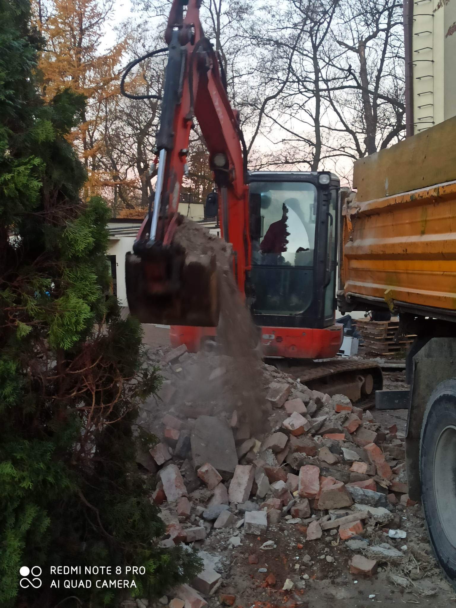 Czerwona mini koparka nabiera gruz z rozbiórki ceglanej ściany i wysypuje do skrzyni żółtej ciężarówki, widoczne drzewa i budynek w tle.