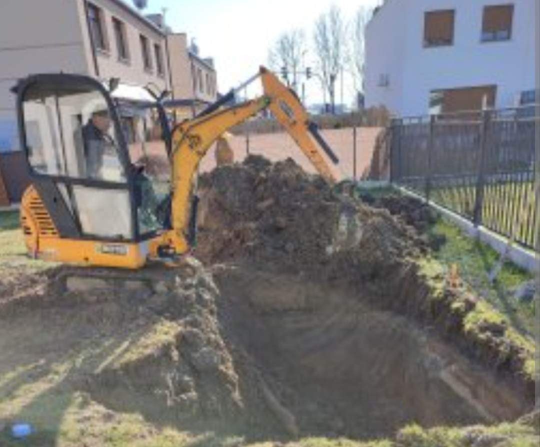 Żółta minikoparka JCB podczas prac ziemnych, wykop pod fundamenty w pobliżu ogrodzenia i budynków mieszkalnych.
