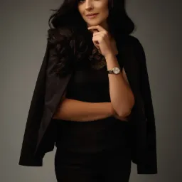 Portret kobiety w czarnym stroju, z marynarką narzuconą na ramiona, w zamyśleniu dotyka brody, na nadgarstku złoty zegarek.