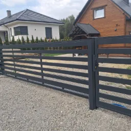 Nowoczesna, metalowa brama wjazdowa w kolorze grafitowym. Horyzontalne, płaskie profile. Domy w tle. Brama zamontowana na posesji z podłożem z kamyków.