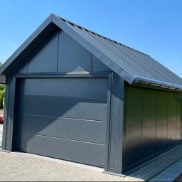 Nikmet Marcin Nikodem - Nowoczesny, grafitowy garaż blaszany z bramą segmentową, na kostce brukowej, z traktorem w tle i widocznym systemem rynnowym.