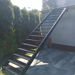 Nikmet Marcin Nikodem - Zewnętrzne, metalowe schody kratowe prowadzące na balkon z balustradą, na tle zielonych drzew i szarej elewacji budynku. Widok z dołu, w słoneczny dzień.