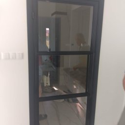 Nikmet Marcin Nikodem - Nowoczesne, czarne drzwi metalowe z szybą, podzielone na trzy sekcje, w jasnym wnętrzu. Widoczne odbicie osoby i wyposażenia.