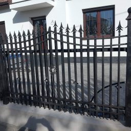 Nikmet Marcin Nikodem - Czarna, metalowa brama wjazdowa z dekoracyjnymi grotami na tle białego domu z brązowymi oknami. Widoczne cieniowanie i kostka brukowa.