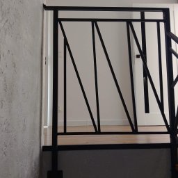 Nikmet Marcin Nikodem - Nowoczesna balustrada schodowa z czarnego metalu o geometrycznym wzorze, widoczna na tle białej ściany i drzwi. Minimalistyczny design, surowe wykończenie ściany.