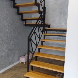 Nikmet Marcin Nikodem - Nowoczesne schody metalowe z drewnianymi stopniami i geometryczną balustradą na tle szarej ściany z fakturą. Widoczne dwa pufy i fragment podłogi.